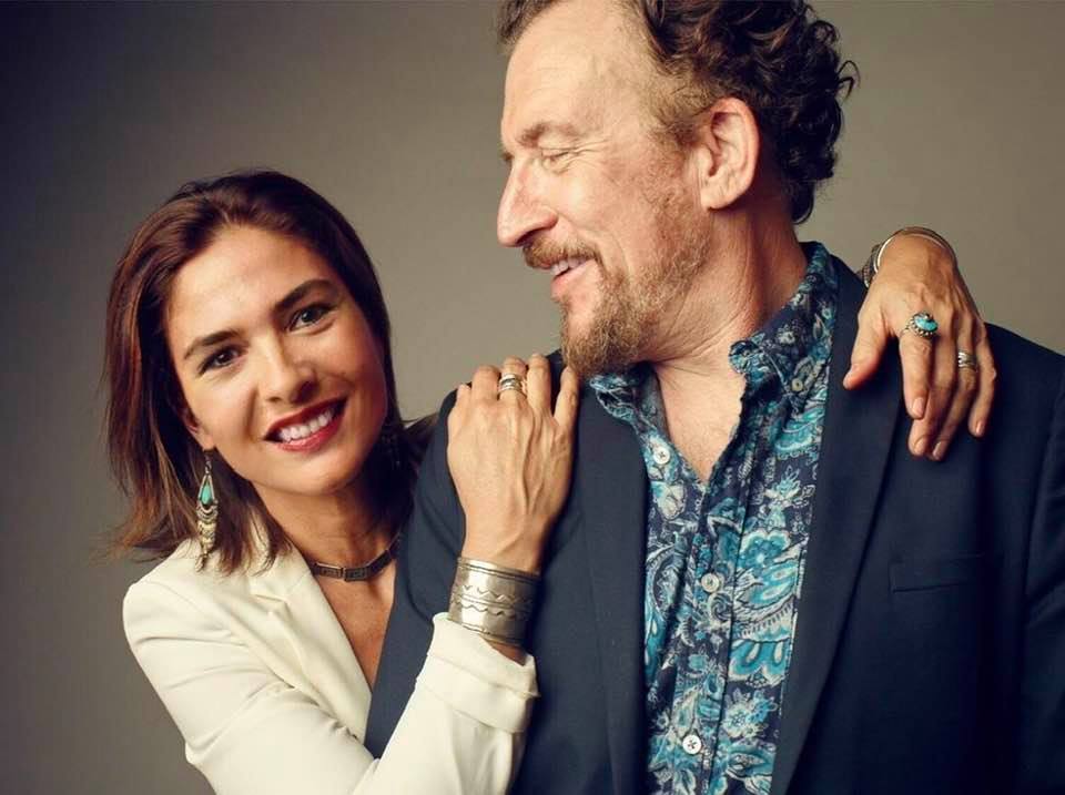 Matthew Asner in a floral print shirt and dark blue coat smiling with his wife n a white dress.